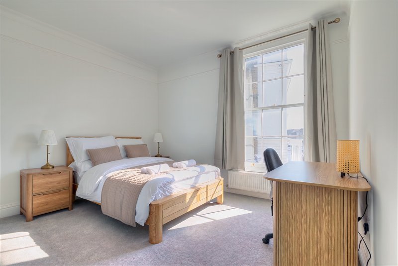 Bright bedroom with wooden bed frame, bedside table, desk, and large window with curtains