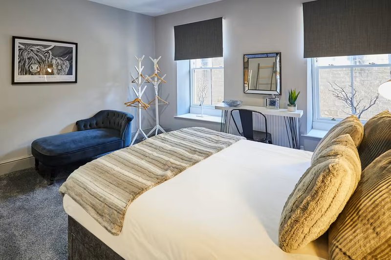 Modern bedroom with double bed, blue chaise lounge, desk area, and large windows with city views