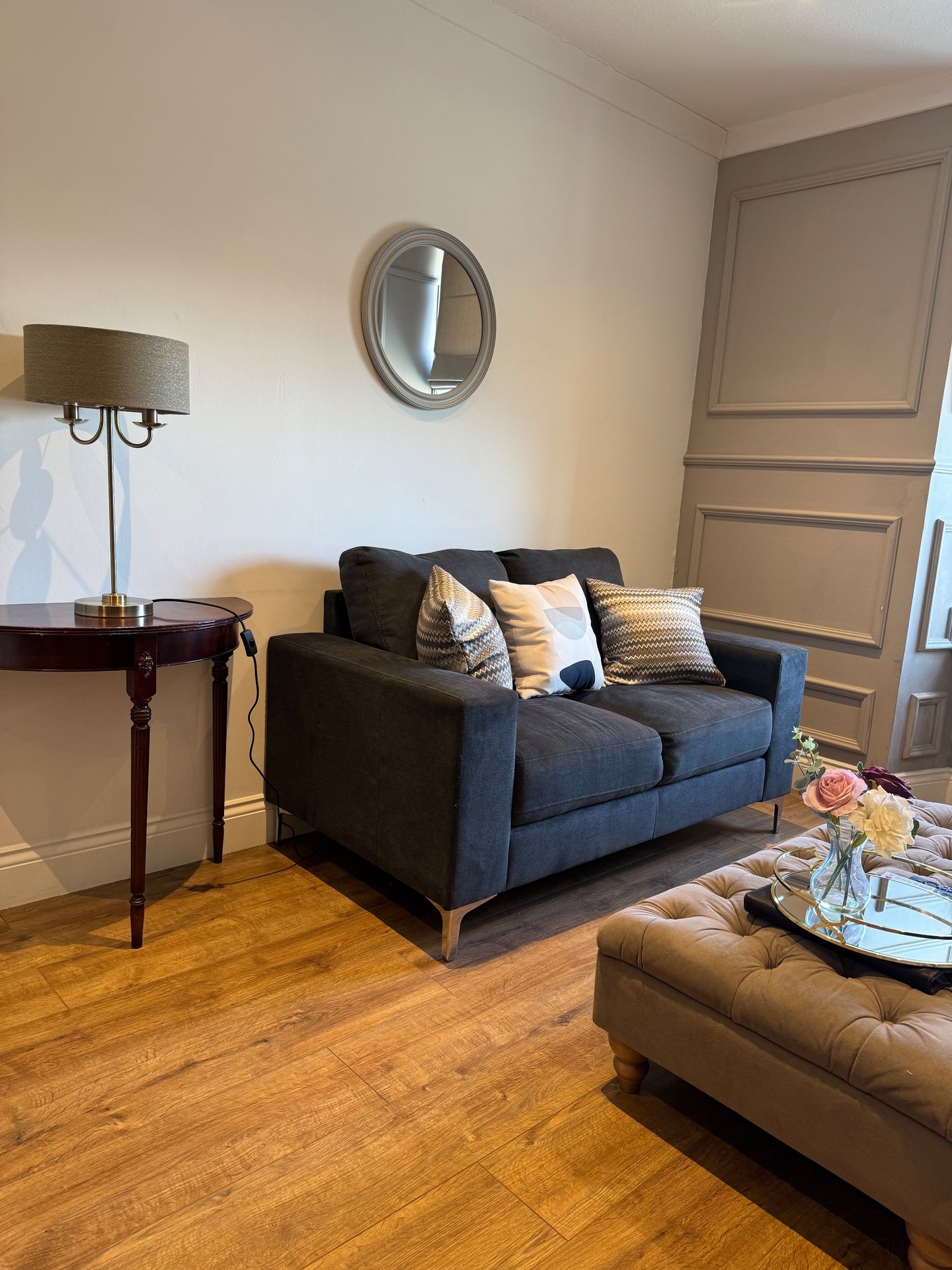 Modern living room with dark blue sofa, wooden side table, round mirror, and wooden flooring