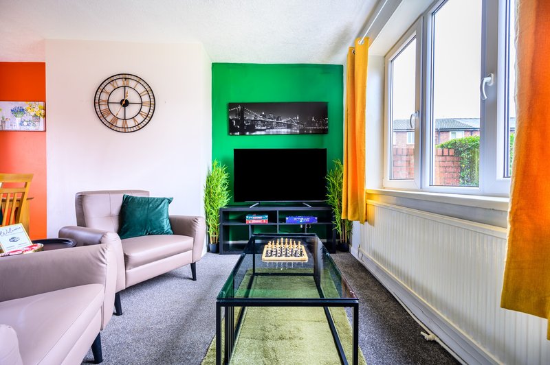 Colorful living room with pink armchairs, wall-mounted TV on green accent wall, clock, and large windows