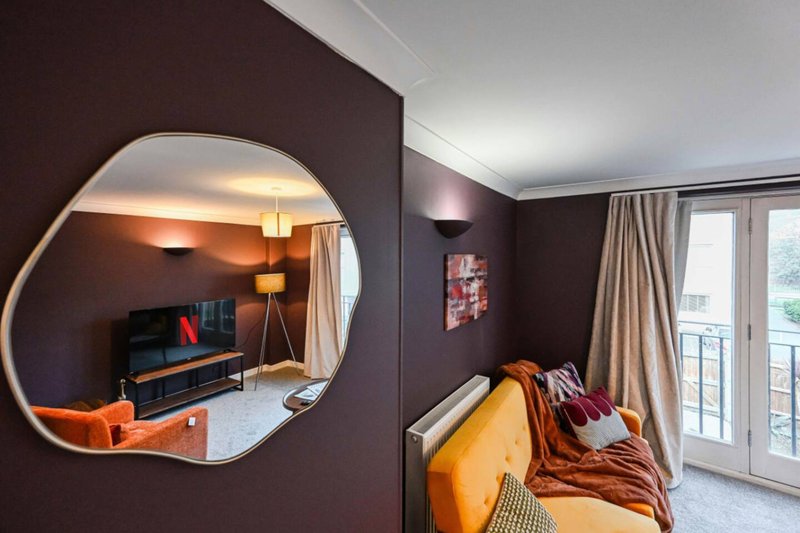 Modern bedroom with dark purple walls, round mirror, desk area, yellow bedding and balcony doors