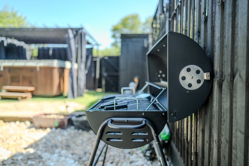 Black charcoal barbecue grill mounted on wooden fence with garden and outbuildings behind