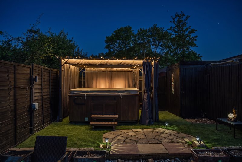 Illuminated hot tub with pergola in enclosed garden at dusk, wooden fencing
