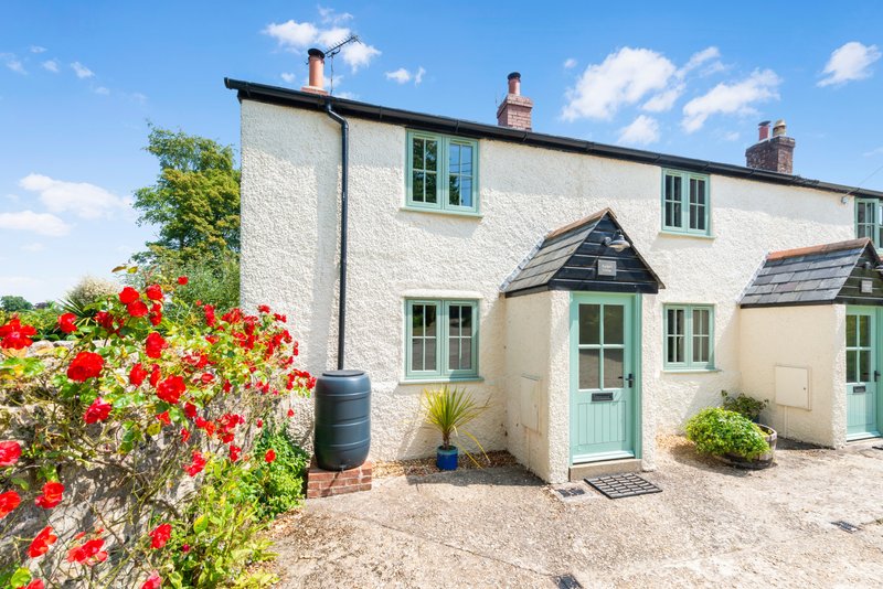 White cottage exterior with green windows, gravel driveway, red flowering shrubs and entrance door