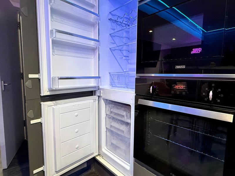 Modern kitchen with open refrigerator, built-in oven and dark cabinetry with blue lighting