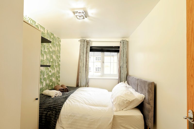 Compact bedroom with double bed, grey headboard, green leaf wallpaper accent wall, and window with curtains