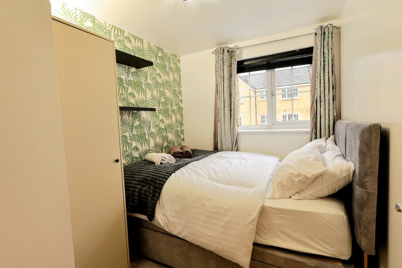 Compact bedroom with double bed, green patterned wallpaper, window with curtains, and cream bedding