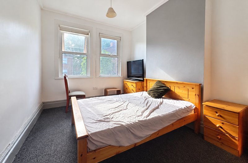 Bright bedroom with wooden bed, white bedding, wooden nightstand, small TV, and window with natural light