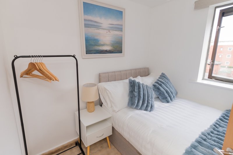 Bright bedroom with double bed, blue striped cushions, wooden hangers on stand, and landscape artwork