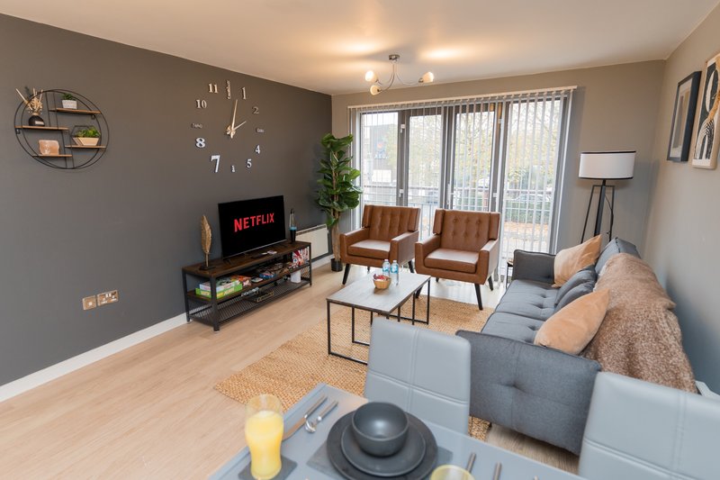 Modern living room with gray sofa, wooden chairs, TV on stand, and large windows
