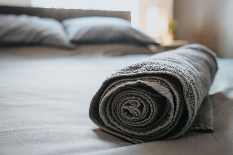 Rolled grey towel on bed with pillows and headboard in background