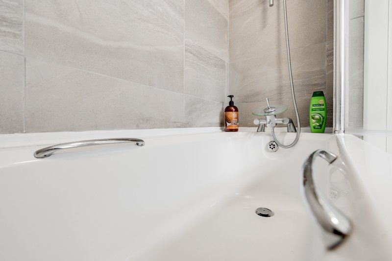 Modern bathroom with white bathtub, gray tile walls, and toiletries on edge