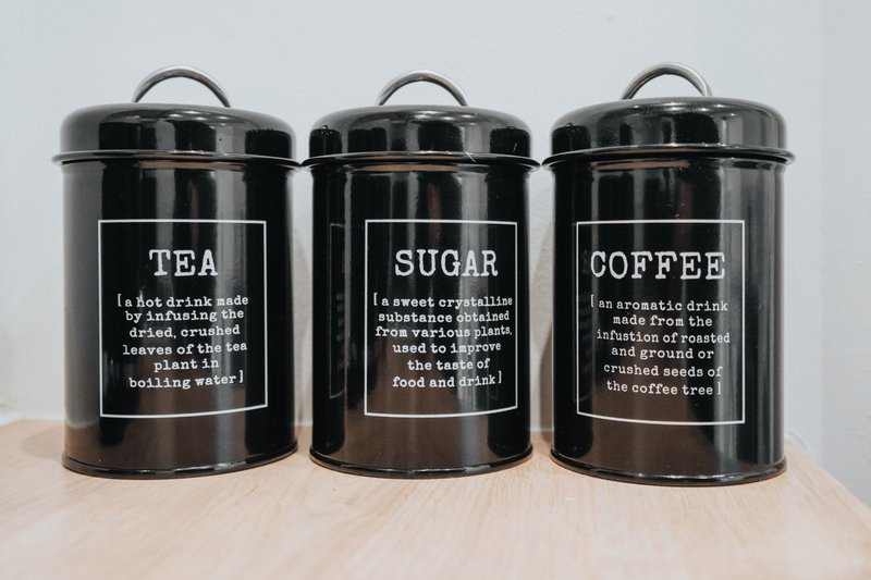 Three black metal kitchen canisters labeled tea, sugar, and coffee on wooden surface
