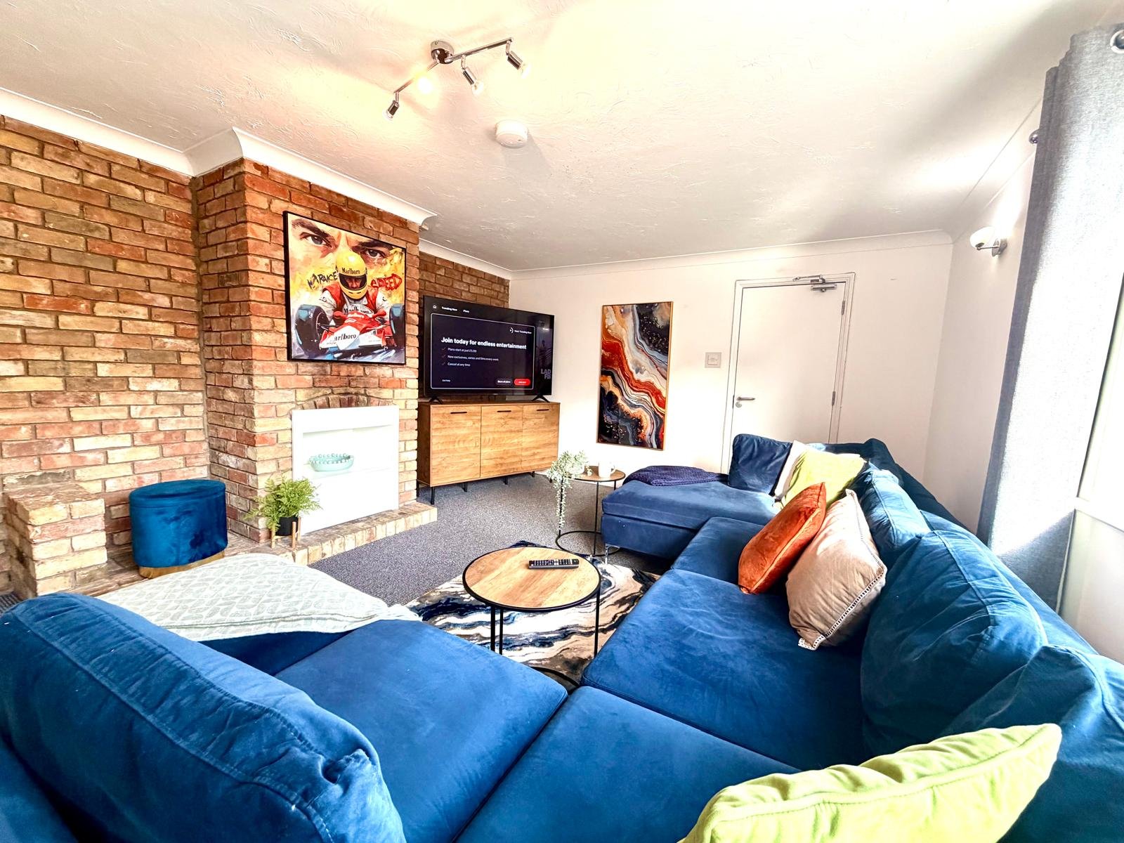 Modern living room with blue L-shaped sofa, brick feature wall, TV, and colorful wall art