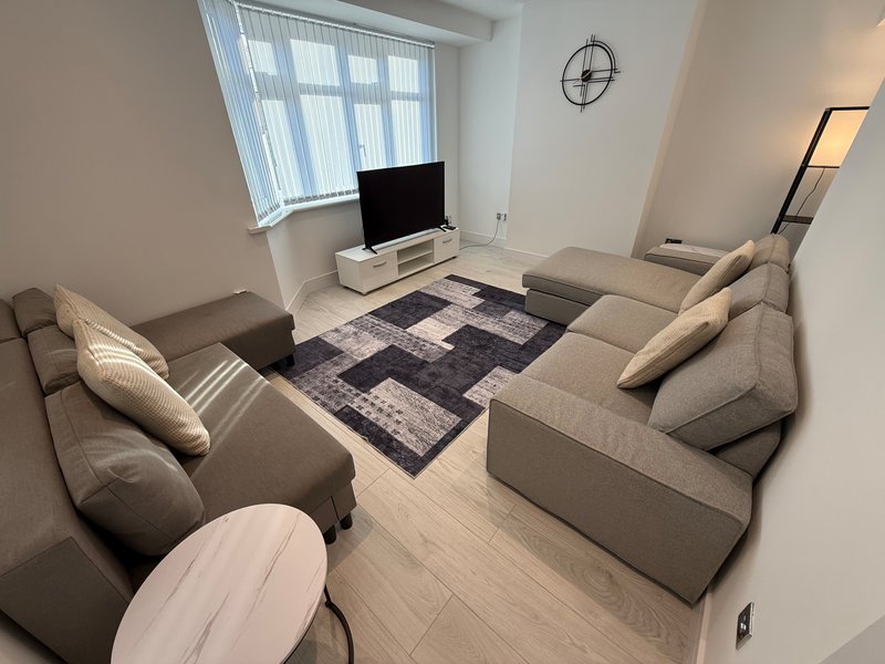 Light living room with grey sofa, TV stand, geometric rug, window with blinds and wall clock