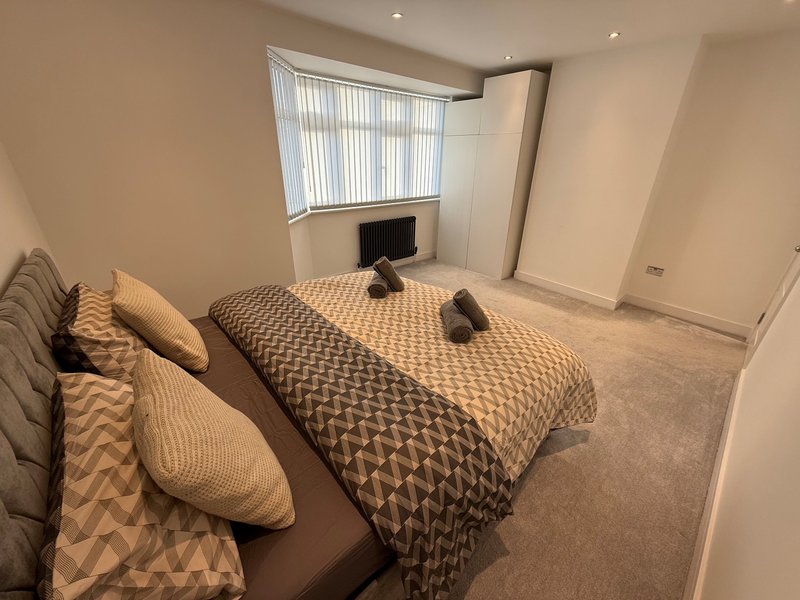 Bedroom with double bed, patterned bedding, cushions, window with vertical blinds and monitor on desk