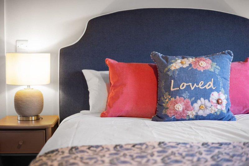Bedroom with dark upholstered headboard, colorful decorative pillows, bedside lamp and wooden nightstand