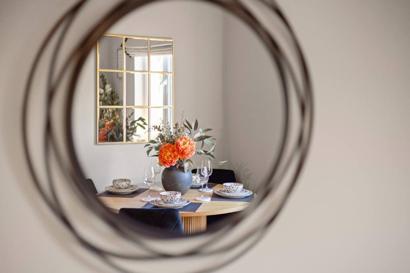 Dining area with round table, blue vase with orange flowers, place settings, and window view through circular mirror frame