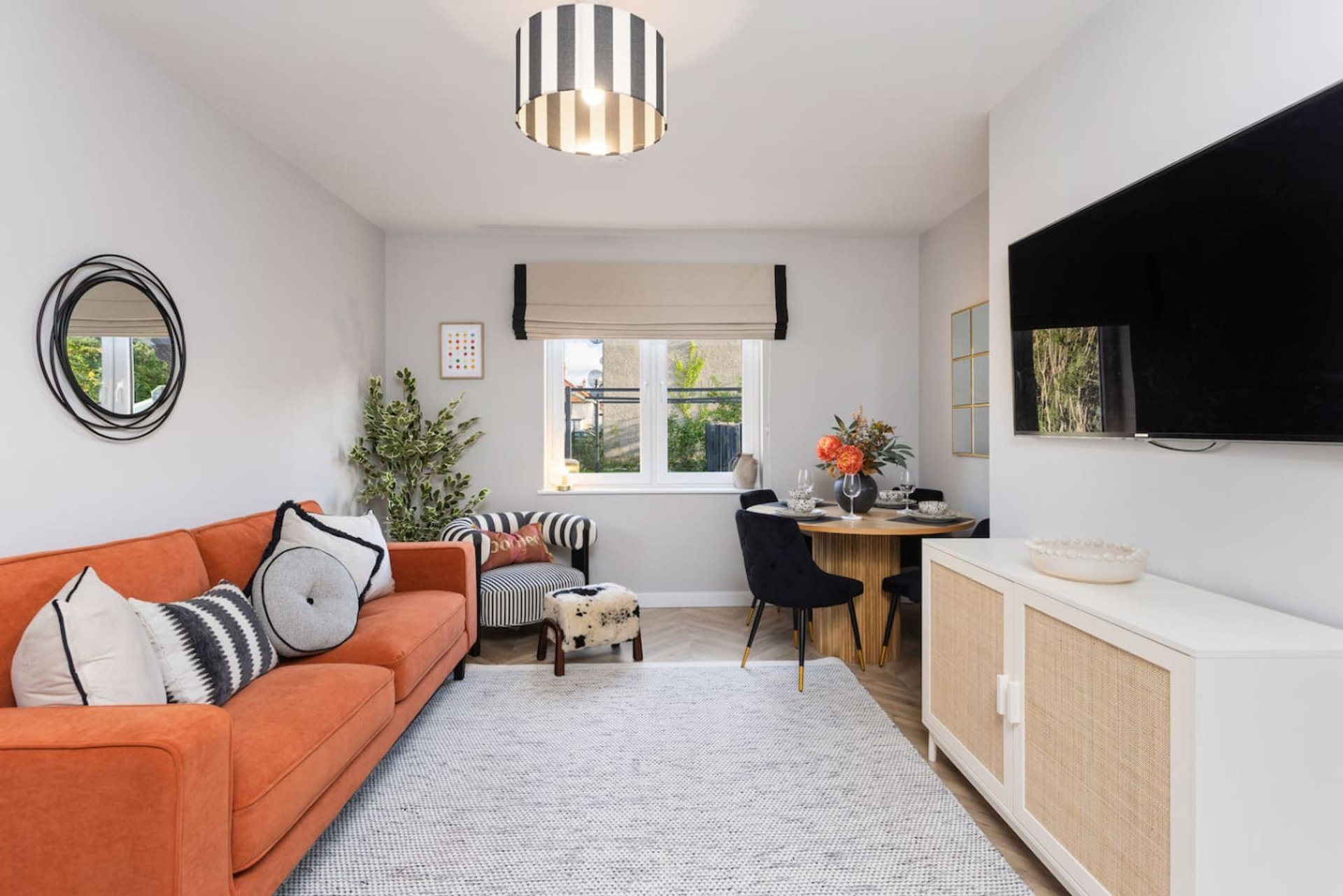 Modern living room with orange sofa, wall-mounted TV, wooden desk, and large windows