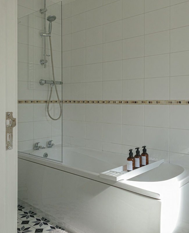 Modern bathroom with white tiles, glass shower screen, bathtub and toiletries on shelf