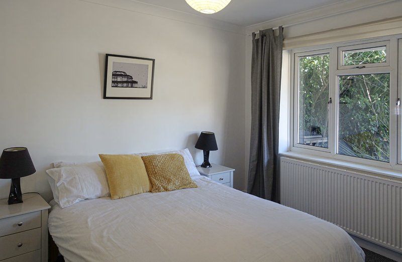 Bright bedroom with double bed, yellow and cream pillows, bedside tables, and large windows with garden views