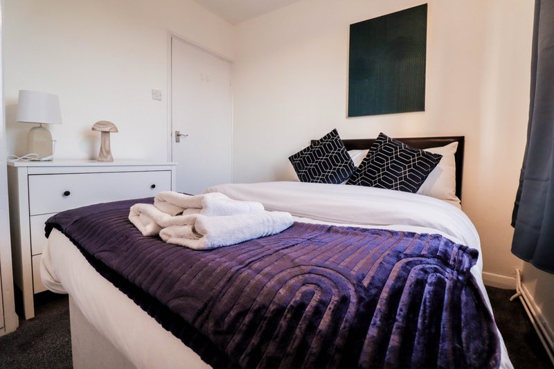 Bedroom with double bed, purple bedding, patterned pillows, white dresser and bedside lamp