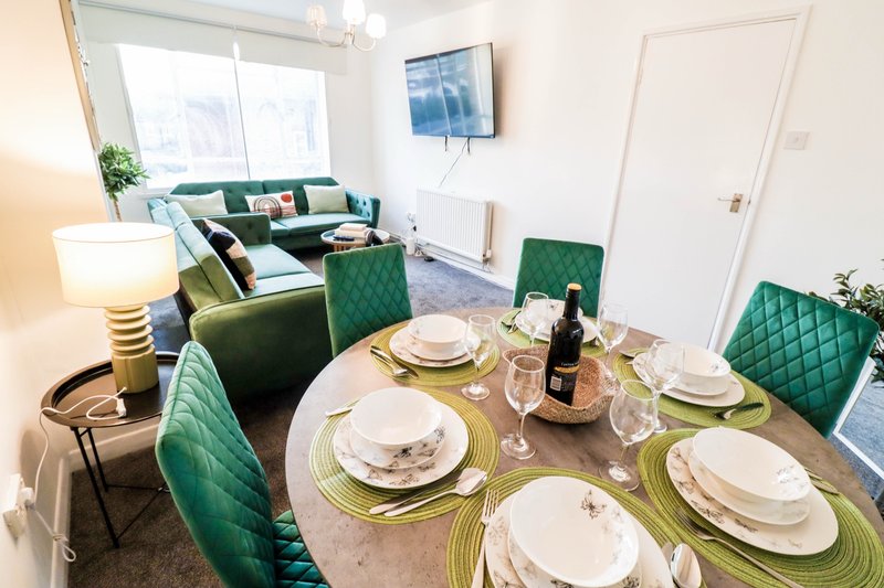Open-plan dining and living area with table set for four, green upholstered chairs, and sofa seating