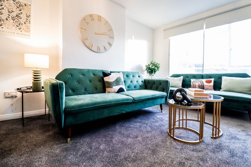 Modern living room with teal velvet sofa, wooden side table, wall clock and bright windows