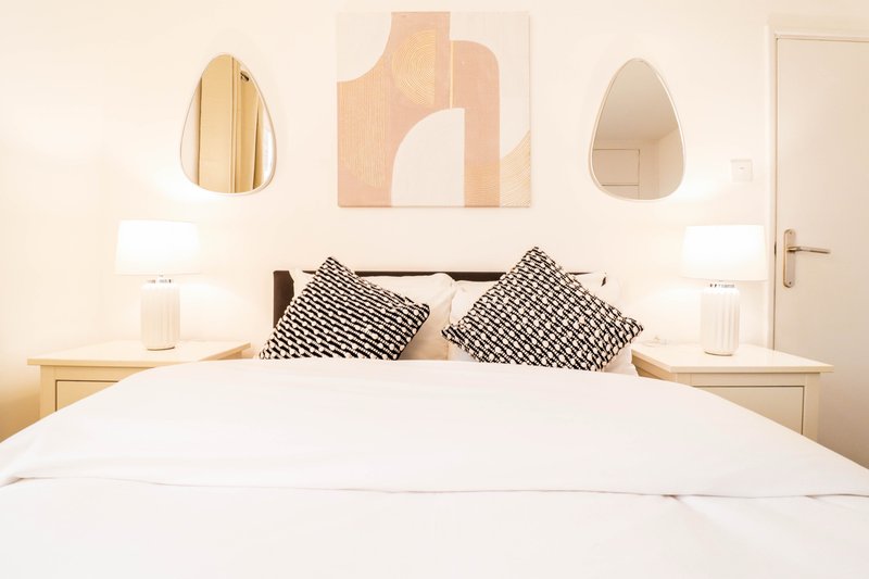 Bright modern bedroom with white bedding, patterned cushions, wooden nightstands, and abstract wall art
