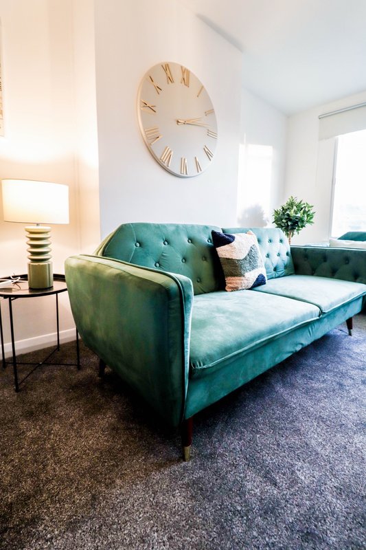 Bright living room with teal velvet sofa, wooden wall clock, side table with lamp and potted plant