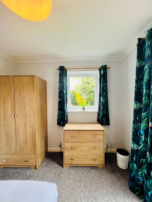 Bright bedroom with wooden wardrobe, chest of drawers, window with dark curtains and green foliage print