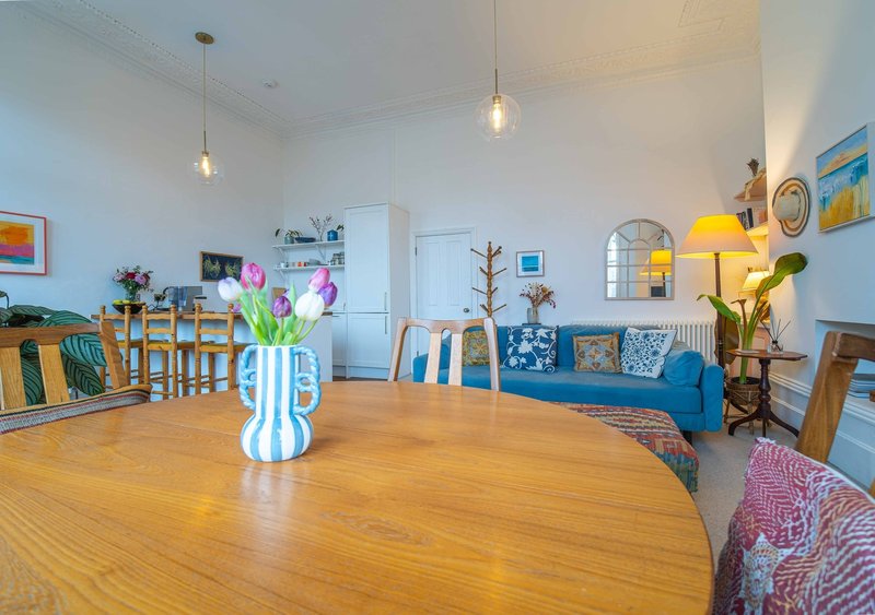 Open-plan living space with wooden dining table, blue sofa, pendant lights, and kitchen area