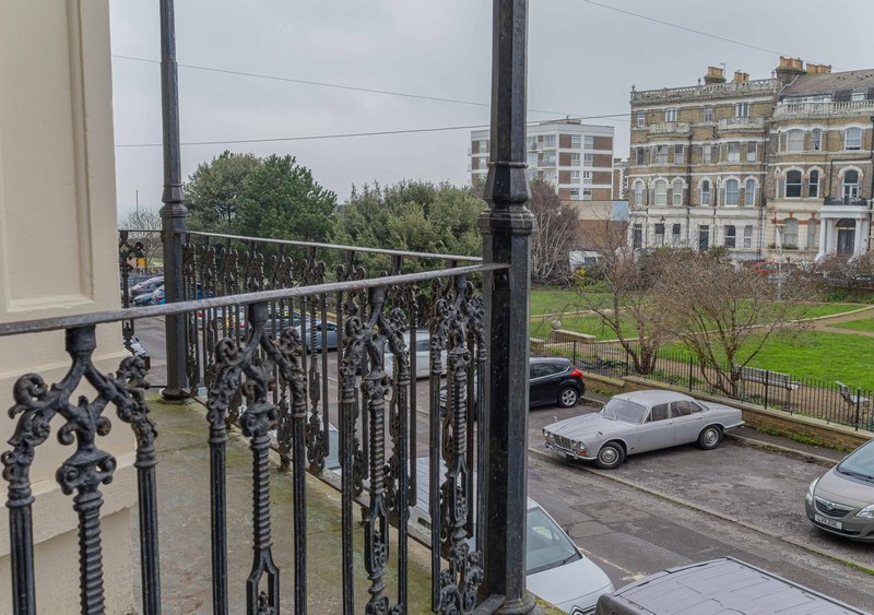 Balcony with ornate black railing overlooking tree-lined square with parked cars and period buildings