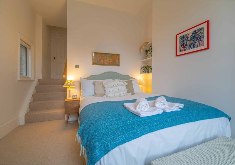 Bright bedroom with double bed, blue throw, bedside lamps, and stairs to upper level