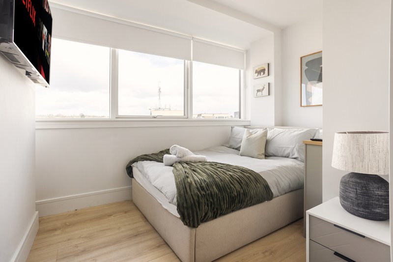 Bright modern bedroom with bed, cushions, wooden floor and large window overlooking city