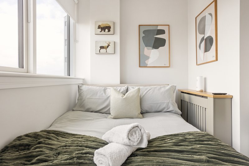 Compact bedroom with double bed, grey and green bedding, cushions, rolled towels, and framed wall art