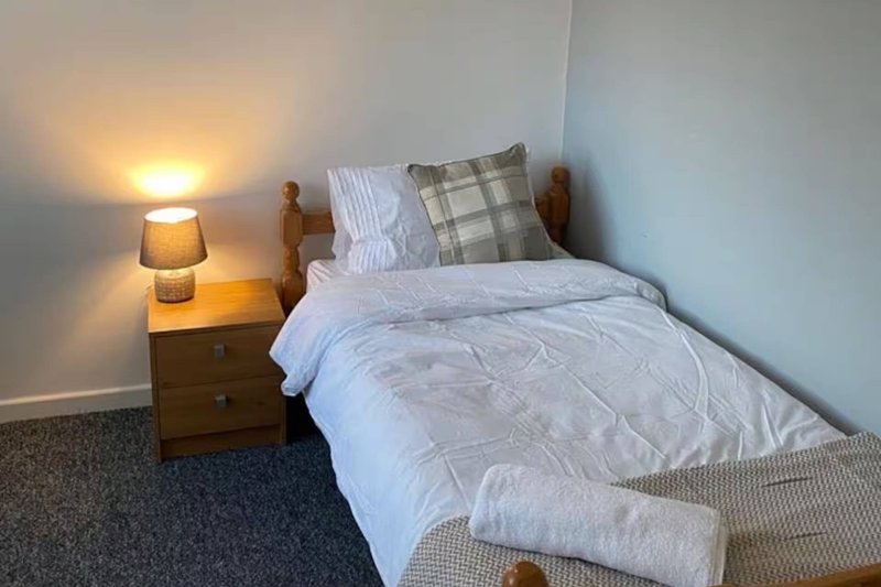 Compact bedroom with single bed, wooden bedside table, lamp, and plaid cushion