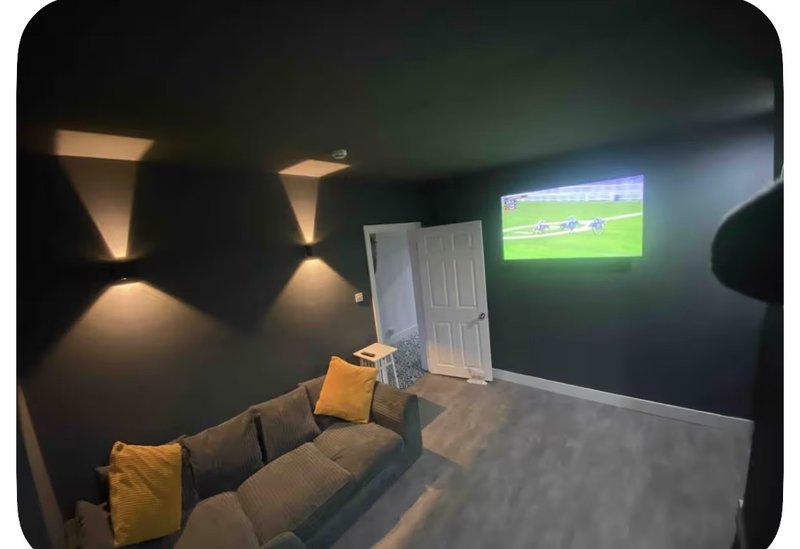 Modern media room with dark walls, wall-mounted lighting, gray sofa with yellow cushions, and wall-mounted TV