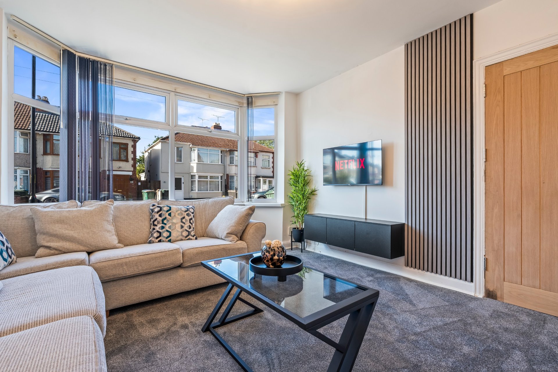 Bright modern living room with beige sectional sofa, black coffee table, TV mounted on wall, large windows overlooking street