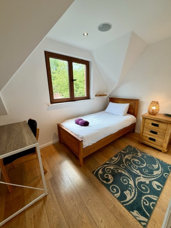 Attic bedroom with wooden bed, window, desk chair, bedside table and patterned floor rug