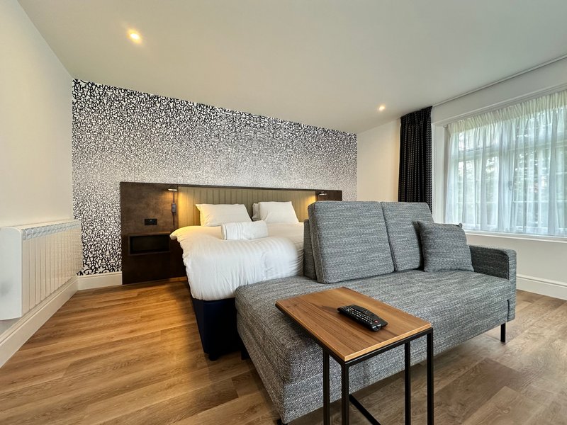 Modern hotel room with grey sofa, wooden coffee table, bed with white linens and textured accent wall