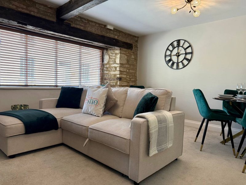 Open-plan living room with cream L-shaped sofa, decorative pillows, wall clock, and dining area with teal chairs