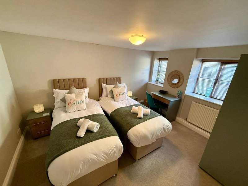 Twin bedroom with two beds, green blankets, white pillows, wooden bedside table, and windows