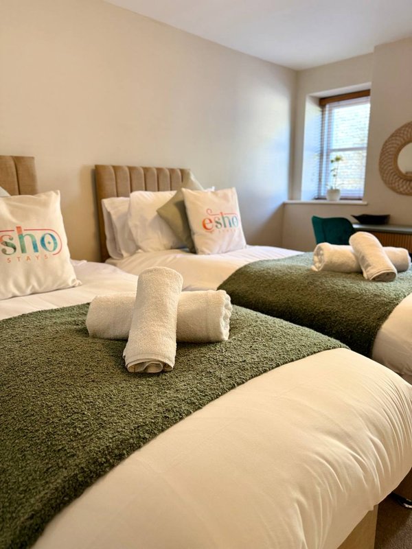 Bright bedroom with two single beds, green throws, rolled towels, and upholstered headboards