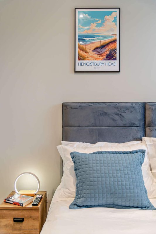 Bedroom with blue headboard, white bedding, blue quilted pillow, wooden nightstand, and coastal artwork on wall