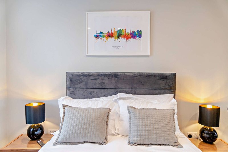 Modern bedroom with grey upholstered headboard, white bedding, decorative pillows, and framed city skyline artwork