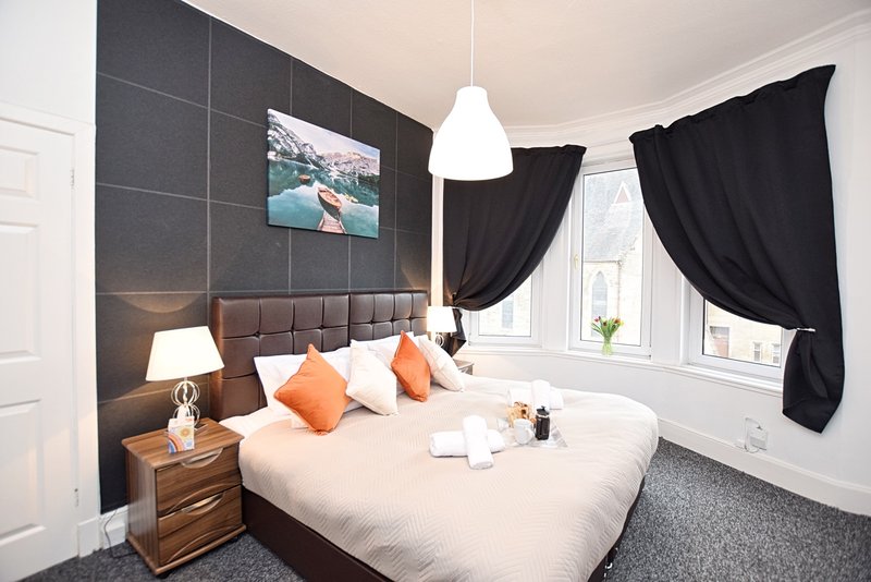 Modern bedroom with king bed, grey upholstered headboard, orange cushions and black curtains
