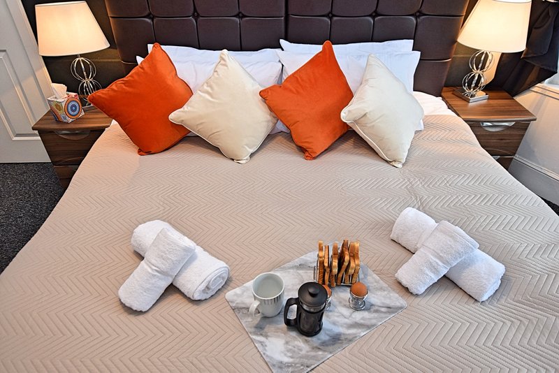Modern bedroom with padded headboard, orange and cream cushions, bedside lamps, and welcome amenities