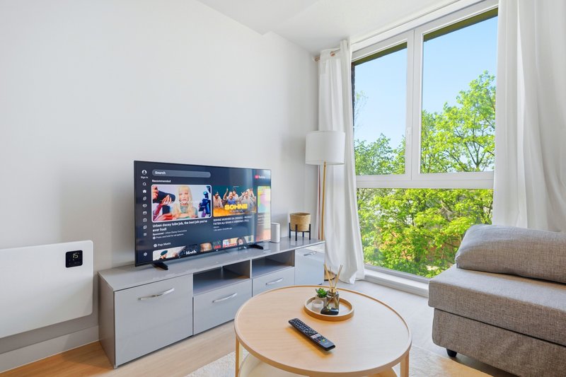 Bright living room with TV, wooden coffee table, gray sofa and large window overlooking green trees