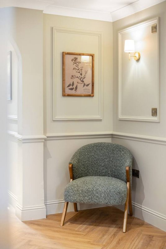 Corner of a light-colored room with an upholstered gray armchair and framed botanical artwork on wall